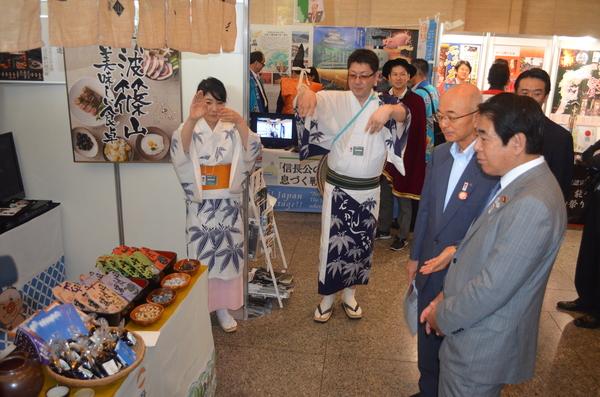 下村博文文部科学大臣が市長と篠山の名産品を見ながらお話をし、隣では浴衣を着た男女が手を上げ踊っている写真