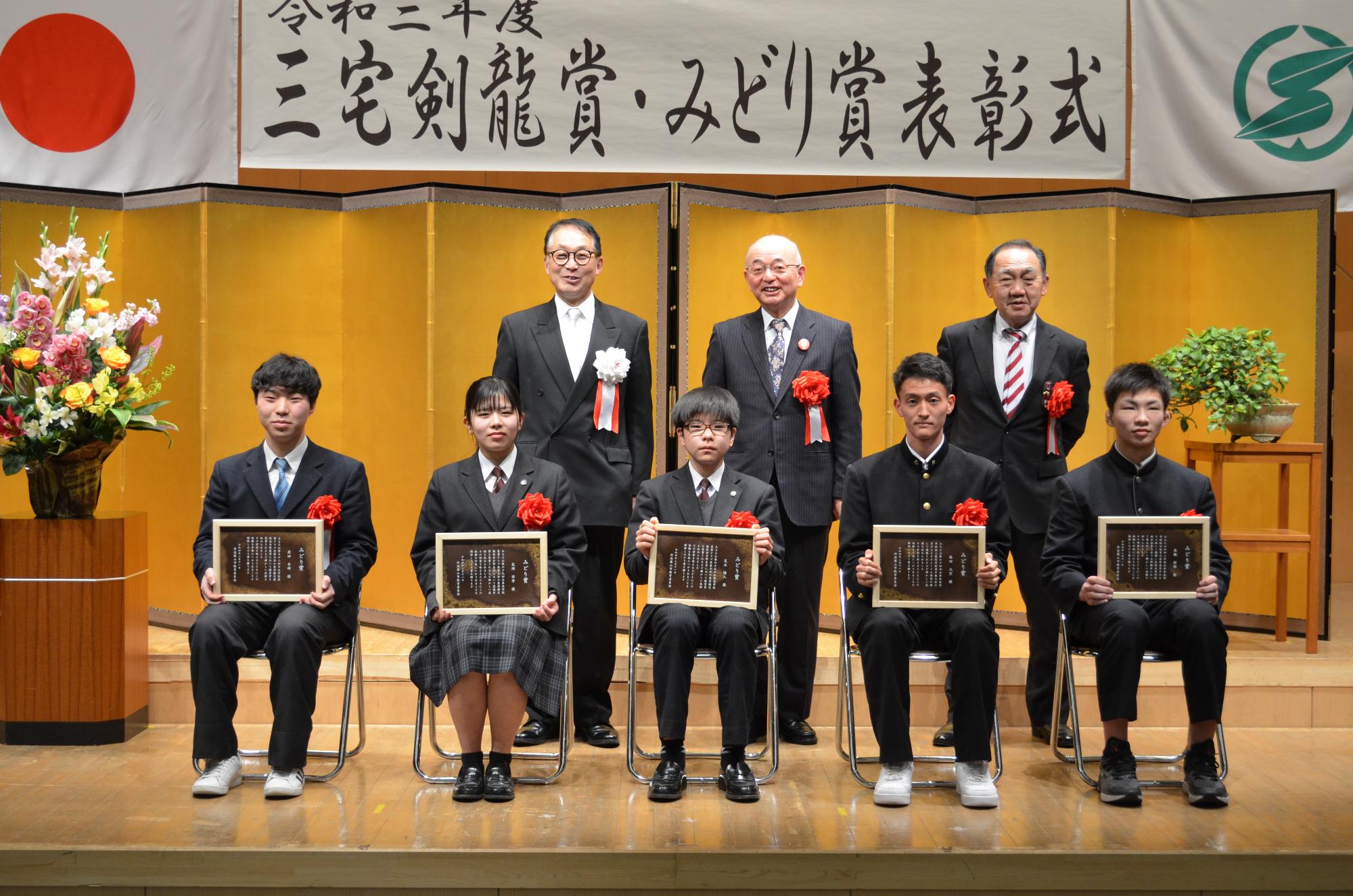 受賞者5名がいすに座って表彰盾をひざにかかげている。後列に市長、教育長、議長が並んでいる