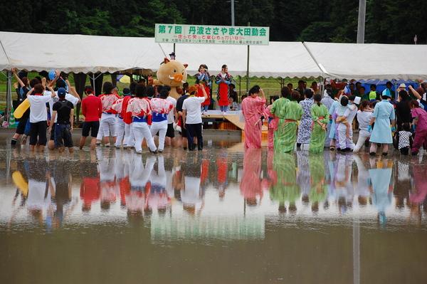 田んぼの中にステージが設置されており、茶娘の衣装を着た女性と、まるいのが立っており、田んぼの中には着物や法被、ナース服など様々な服装の人々がいる写真