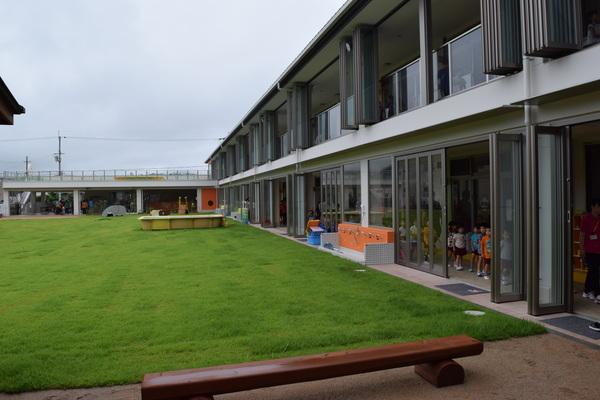 中央に緑の芝の園庭があり、その横に壁がすべて透明のガラスになっている子ども園の建物が立っている写真