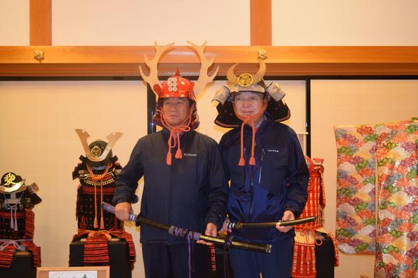 鎧兜や着物が飾られている和室で、鎧兜を被った男性2名が刀を持って記念撮影している写真
