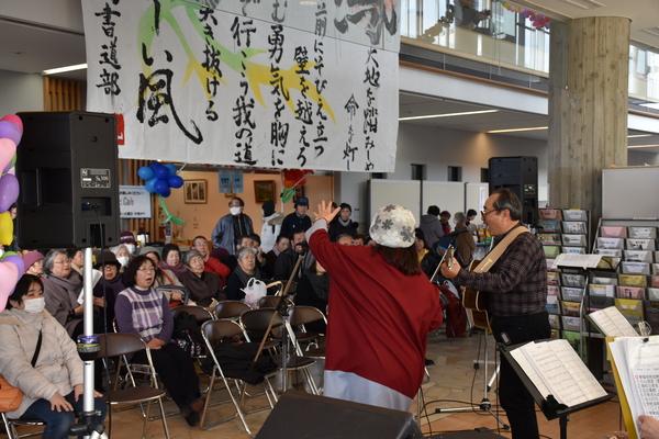 ステージでギターの弾き語りをしている男性と花柄のベレー帽をかぶった女性が手をあげて歌っているのを観客が聴いている様子