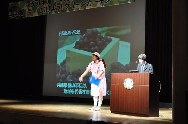 壇上のスクリーンの前で、赤いランドセルと黄色い帽子を被った小学生の恰好をした大人が篠山特産の「丹波黒大豆」について説明しており、右端で司会者が聞いている写真