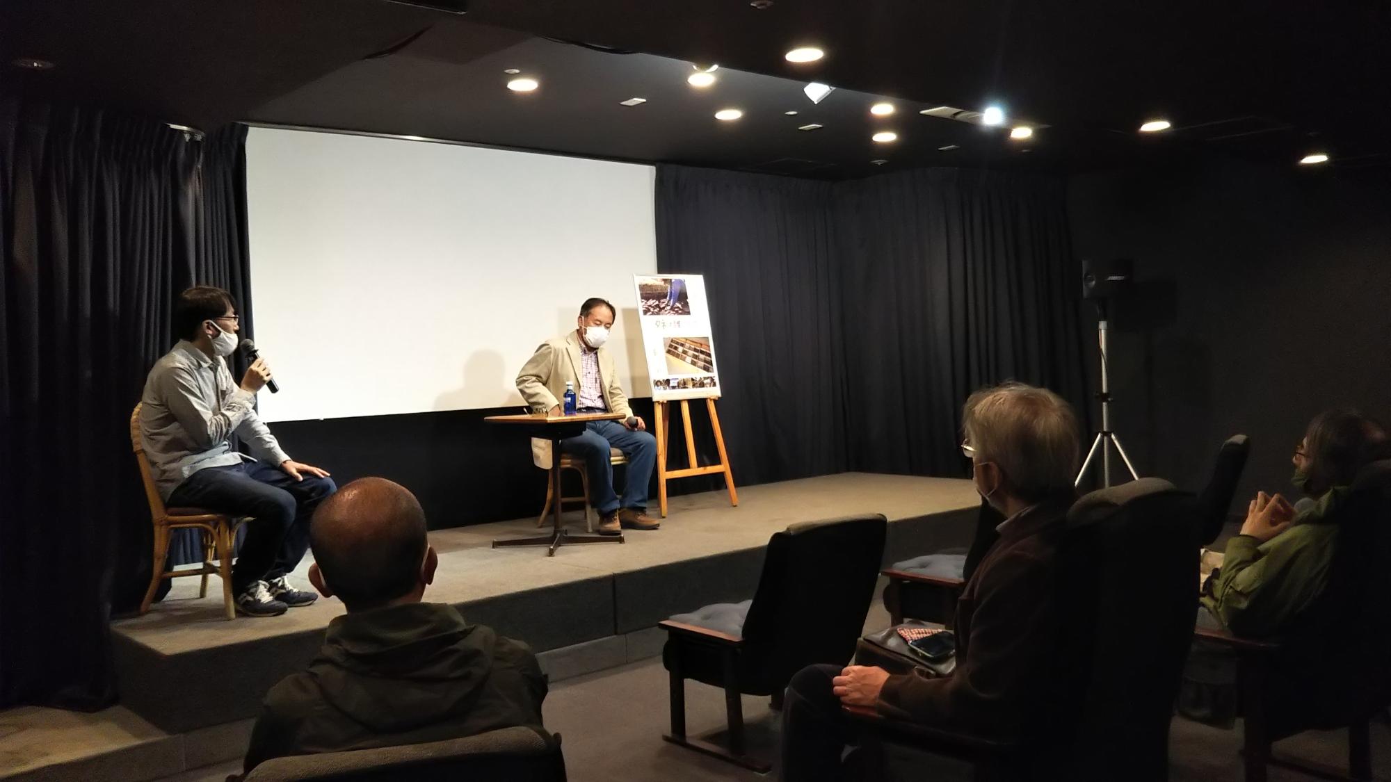 ミニシアターで講師の方が壇上の椅子に座ってパネルを用いて説明されている。進行の方は下手で椅子に座ってマイクでしゃべっている。