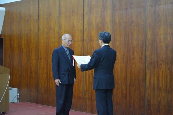 スーツに胸章リボンをつけた男性が、賞状を読み上げる男性の前に立っている写真