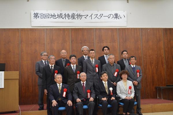 地域特産物マイスタに認定された方々がスーツに胸章リボンをつけ3列に並んで集合写真