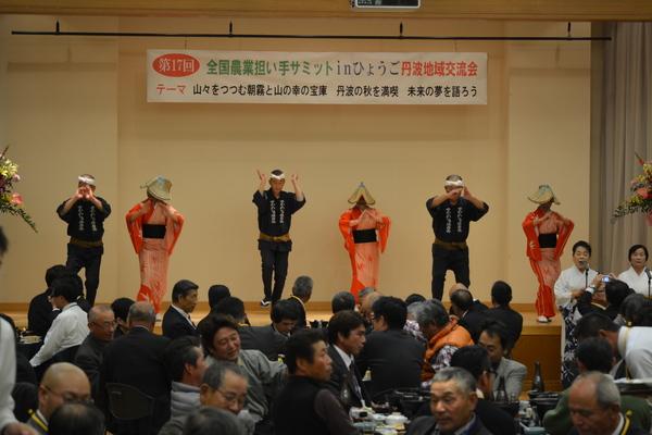 「全国農業担い手サミットinひょうご 丹波地域交流会」と書かれている横断幕の前の舞台上に紺色の半纏と股引きを着ている男性と編み笠をがぶってオレンジ色の着物を着た女性が踊っている様子の写真