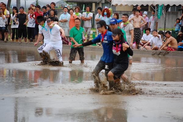 目隠しをして田んぼの中で競争をしている男女とその様子を見ている方々の写真