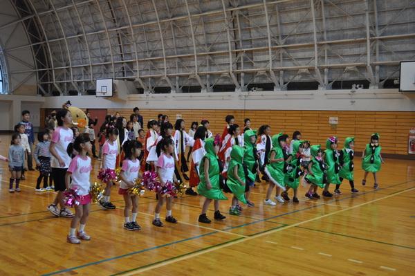 緑の衣装を着た子供達とボンボンを手に持っている女の子が前に並び、その後ろで私服を着た子供達が踊っている写真