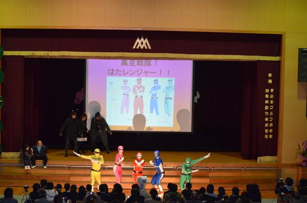 体育館のステージのスクリーンに映し出されている黒豆戦隊はたレンジャーをバックに本物の「黒豆戦隊はたレンジャー」が登場しスポットライトを浴びて決めポーズをしているのを人々が見ている写真