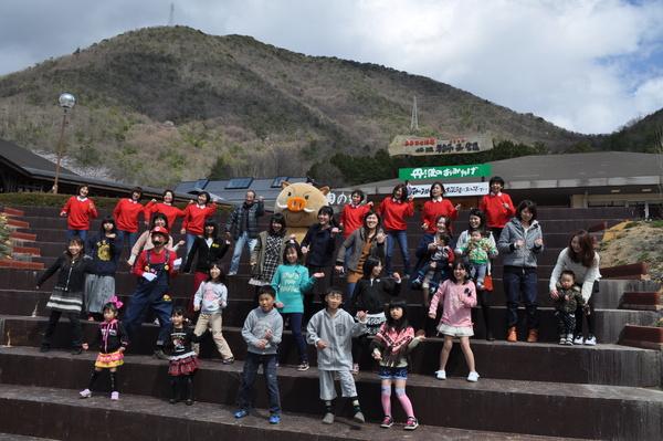 後ろの山をバックに階段で子供達とまるいの、赤いTシャツをきた大人の人達が踊っている写真