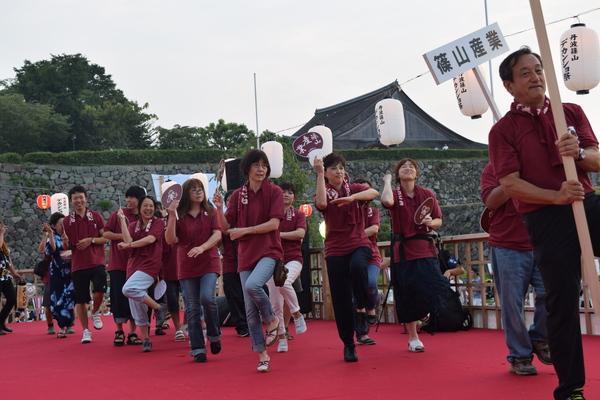 篠山産業のプラカードの後ろを小豆色のポロシャツを着た女性がうちわを持って踊っている写真