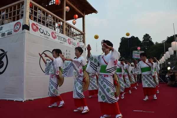 舞台下にひかれた赤い敷物の上で丹南音頭保存会の方が3列に並び浴衣姿で手を上にハの字であげ踊っている写真