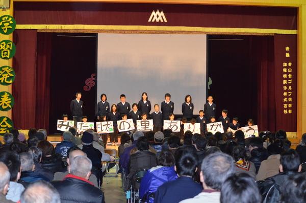 体育館のステージで生徒たちが3列に並んで立っており、最前列の生徒たちが一文字ずつ書かれた画用紙を持って写っている写真