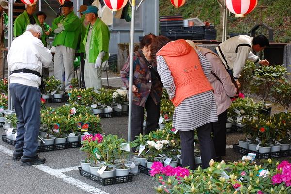 テントの販売ブースでシャクナゲの苗を見ている来場者と奥に法被を着た主催者が集まっている写真