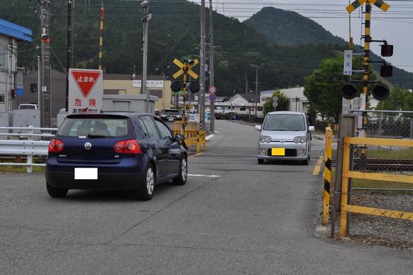 道幅の狭い弁天踏切に手前には青い乗用車、対向車線にはグレーの軽自動車が踏切の停止線で停止している様子の写真