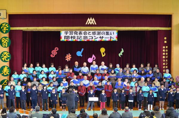 生徒らが畑地区の「みたけの里うぐいす」と「芦屋夢クラブ」のメンバーと一緒に体育館のステージで指揮に合わせて合唱しているのを人々が見ている写真