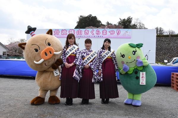 さくらまつりのステージをバックに袴姿の観光大使の女性3名と両脇にゆるキャラが写っている記念写真