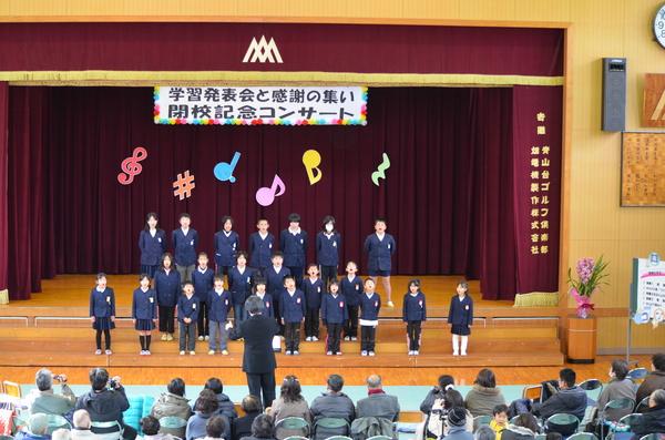 体育館のステージで生徒らが先生の指揮に合わせて合唱しているのを人々が見ている写真