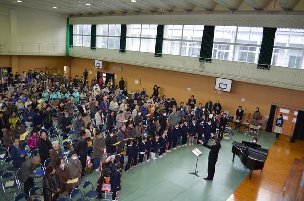 体育館の客席側全員でピアノの演奏と指揮に合わせて歌っている写真