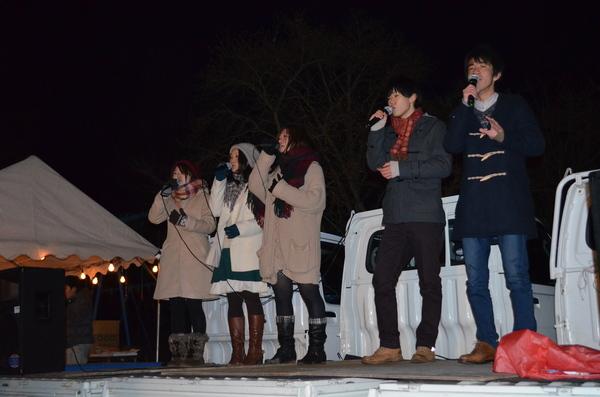 マフラーを首に巻いて、コートを着た3人の女性と2人の男性がトラックの荷台でアカペラを歌っている写真