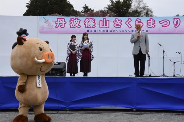 さくらまつりのステージで市長が挨拶しているのをステージ横で見ている袴姿の女性らとステージ下でゆるキャラ（まるいの）が見ている写真