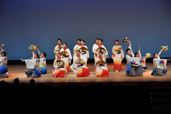 裾に青色の模様が入った衣装を着た女性が金色のボンボンを持ってポースをとって歌を歌っており、裾にオレンジ色が入った衣装を着た女性が金色のセンスを広げ中央で歌を歌っている様子の写真