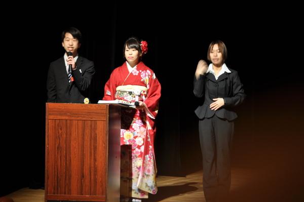 司会の中西君と小倉さんが演台の前に立っており、中西君がマイクを持って話をしている横で手話通訳者の女性が同時通訳している様子の写真