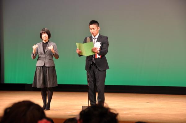 舞台上のスタンドマイクの前で松本君が黄緑色の表紙の紙を広げ話をしており、横に立っている通訳者の女性が同時通訳をしている様子の写真