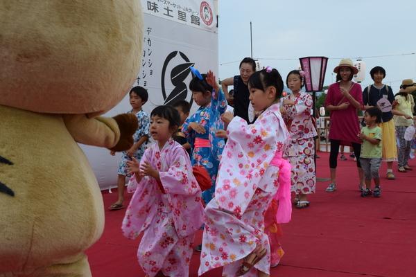 浴衣姿の子供たちや家族連れがゆるキャラ「まるいの」の踊りの動きを見て片足を上げて踊っている写真