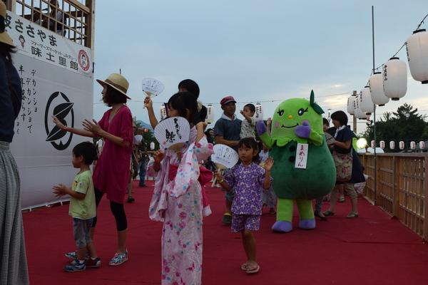 うちわを持った浴衣姿の子供たちや家族連れがゆるキャラ「まめりん」と一緒に提灯が飾ってあるヤグラで踊っている写真