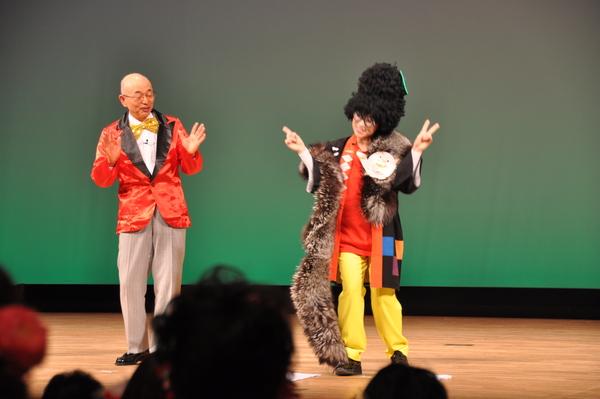 舞台上で赤いタキシードを着た市長と、頭にもじゃもじゃの髪の毛のかつらを被ってメガネを掛け、首に毛皮のようなものをかけ、「まるいの」の顔のイラストが貼られた衣装を着けた女性がビースサインをしており、それを見て市長がびっくりしている様子の写真