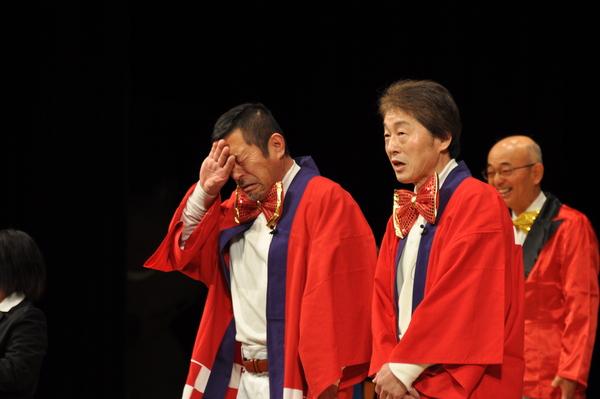 赤い蝶ネクタイを付けて赤い法被を着た男性2名が舞台上に立っており、左側の男性が泣いている様な仕草をしており、後にいる市長が笑っている様子の写真