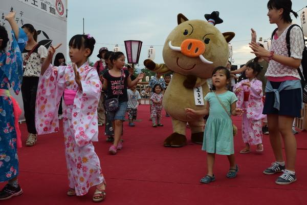 浴衣姿の子供たちや家族連れと一緒にゆるキャラ「まるいの」も交じって両手を挙げて踊っている写真