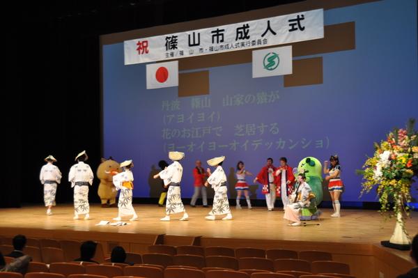 網傘を被り笹の模様が入った浴衣を着ている踊り子とまるいの、まめりん、市長などが舞台上で円になってデカンショを踊っており、舞台のスクリーンに歌の歌詞写っている様子の写真