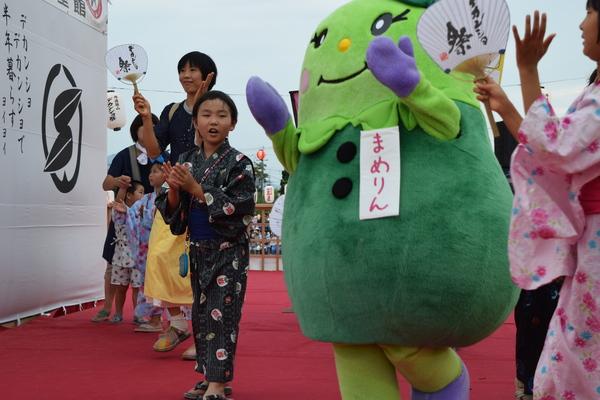 浴衣姿の子供たちや家族連れがゆるキャラ「まめりん」の動きを真似しながら手をたたいて一緒に踊っている写真