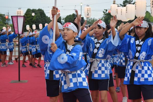 プラカードを持った男の子を先頭に鉢巻きと青い法被姿の子供らが両手を挙げて踊っている写真