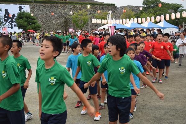 緑・水色・紫・赤色のTシャツを着た子供たちが両腕を広げて踊っている写真