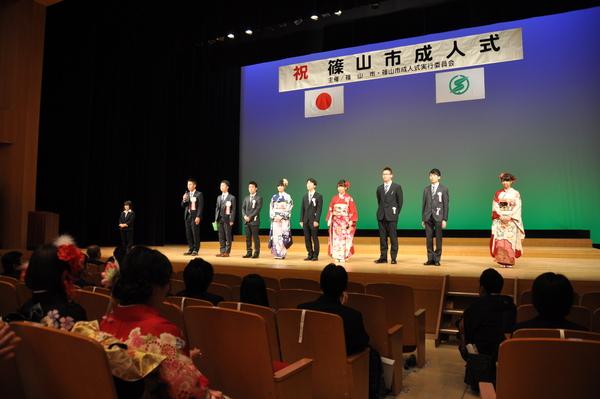 舞台の上に、スーツを着た新成人が6名、振袖を着た新成人3名立っており、左奥のスーツを着た男性がマイクを持って話をしている様子の写真