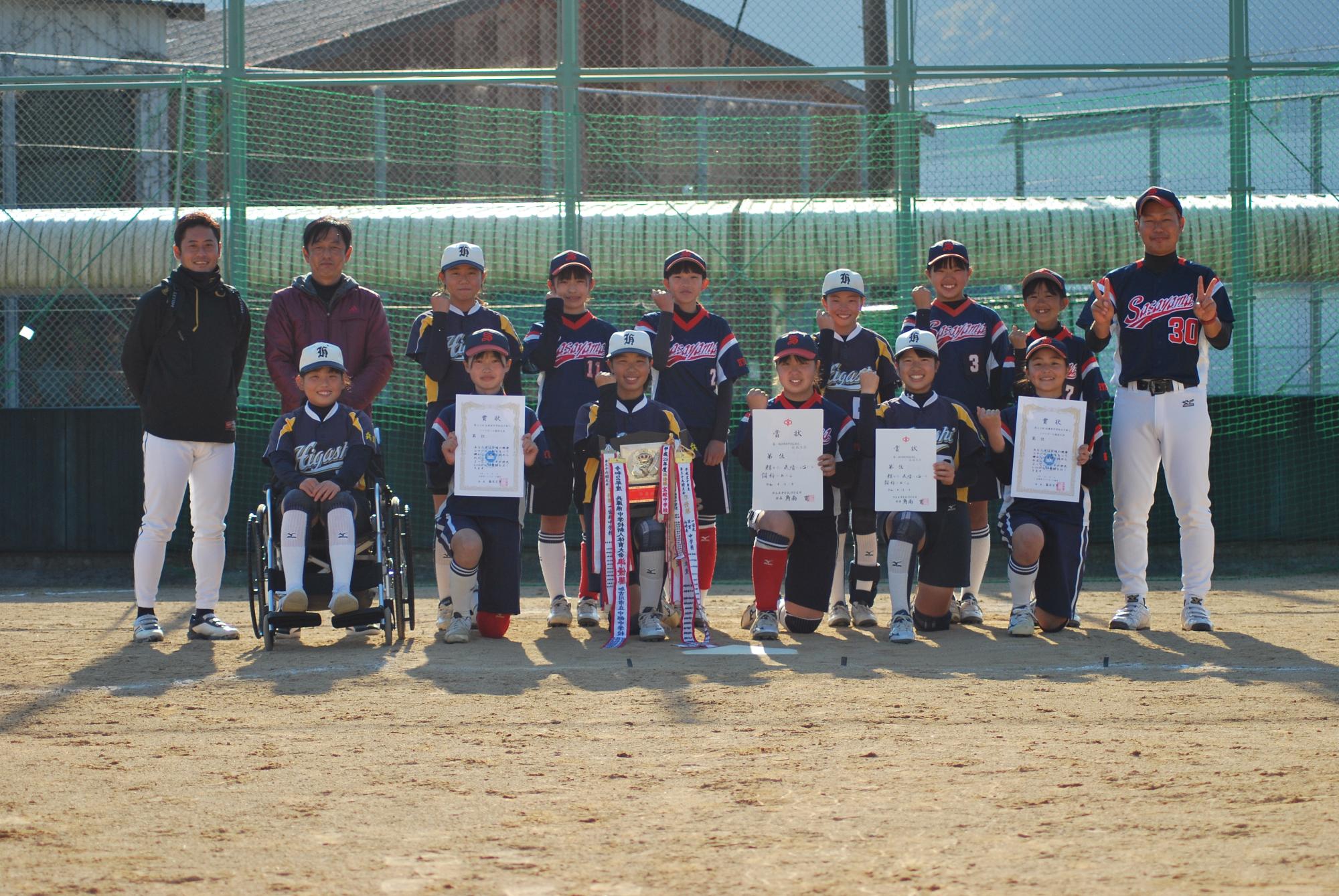 ソフトボールの試合で準優勝し、盾や表彰状を持って部員と顧問の先生たちが記念撮影している