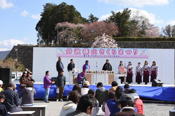 さくらまつりのステージでビンゴゲーム大会が行われている様子と参加している人々の写真