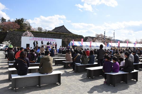 さくらまつりのステージのイベントを椅子に座って観覧している人々と、ステージ横のテントやのぼり旗が写っている写真