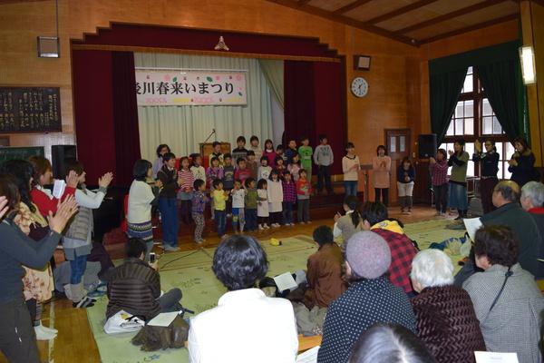 体育館の前方で後川小学校の校歌を斉唱している子供たちと拍手をしている観客たちの写真