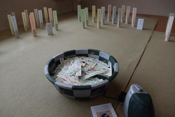 器の中にスカート、鉛筆など文字の描かれた紙がたくさん入っている作品が置かれている写真