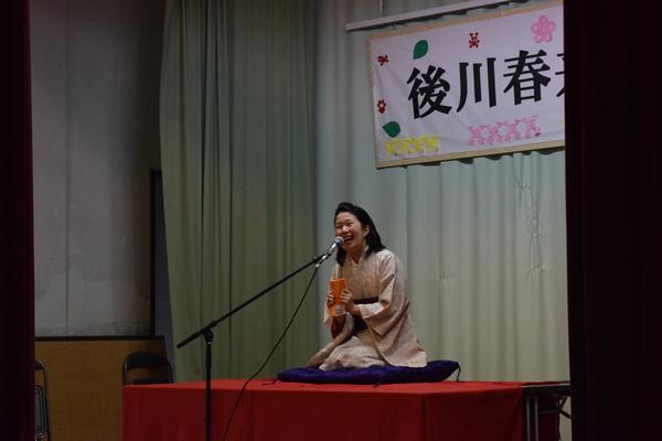 着物を着た島井さんがステージの上で座布団の上に正座で座り笑顔で落語の話をしている写真