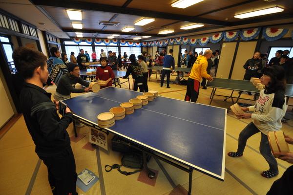 卓球のコートの中央にネットの代わりに桶が置かれていて、参加者がラケットの代わりに桶を持って卓球をしている写真