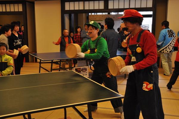 マリオのコスプレをした男性とルイージのコスプレをした女性が桶を持って卓球をしている写真