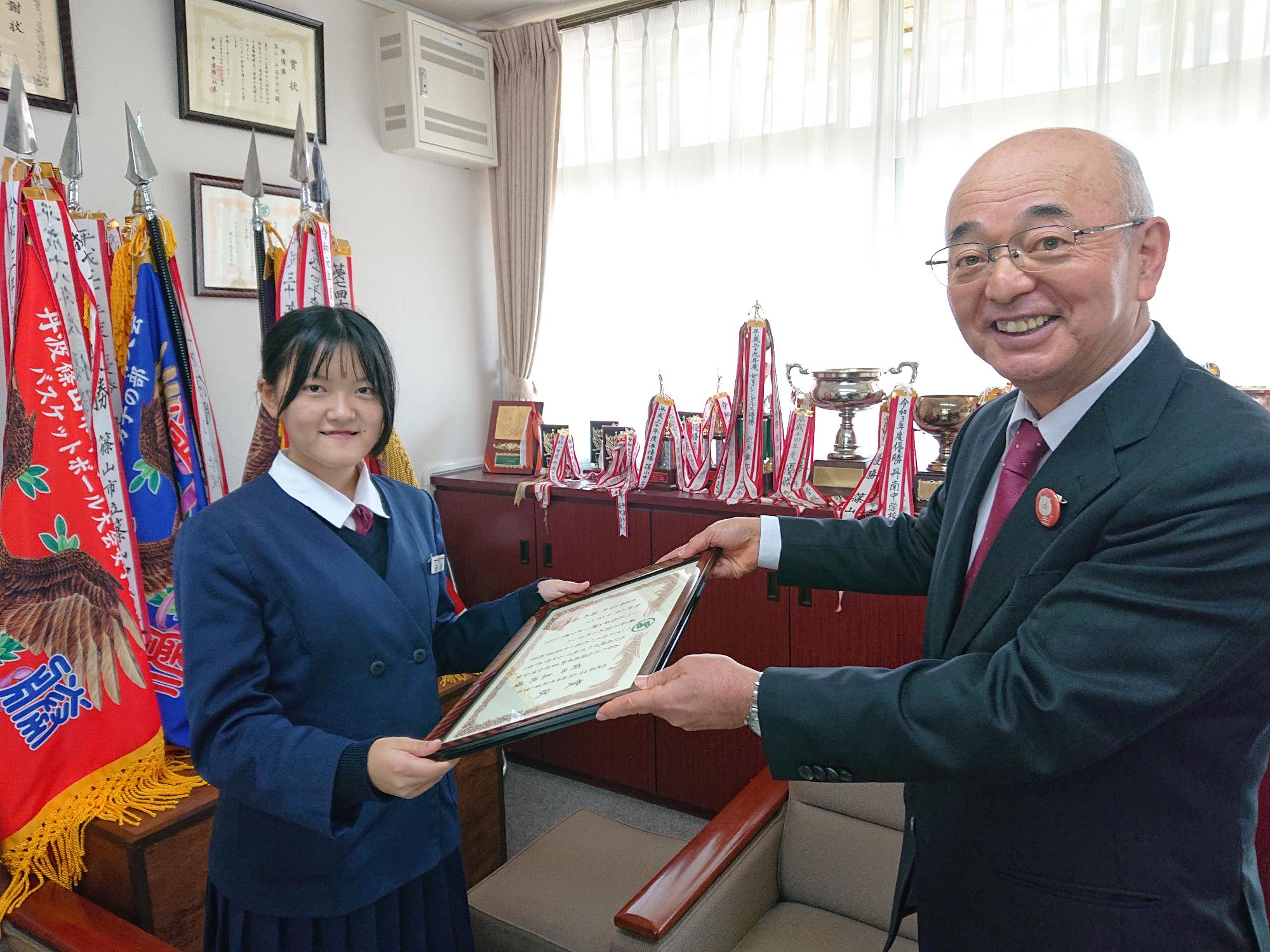 制服を着た女の子が、市長から額に入った表彰状を受け取ろうとしている