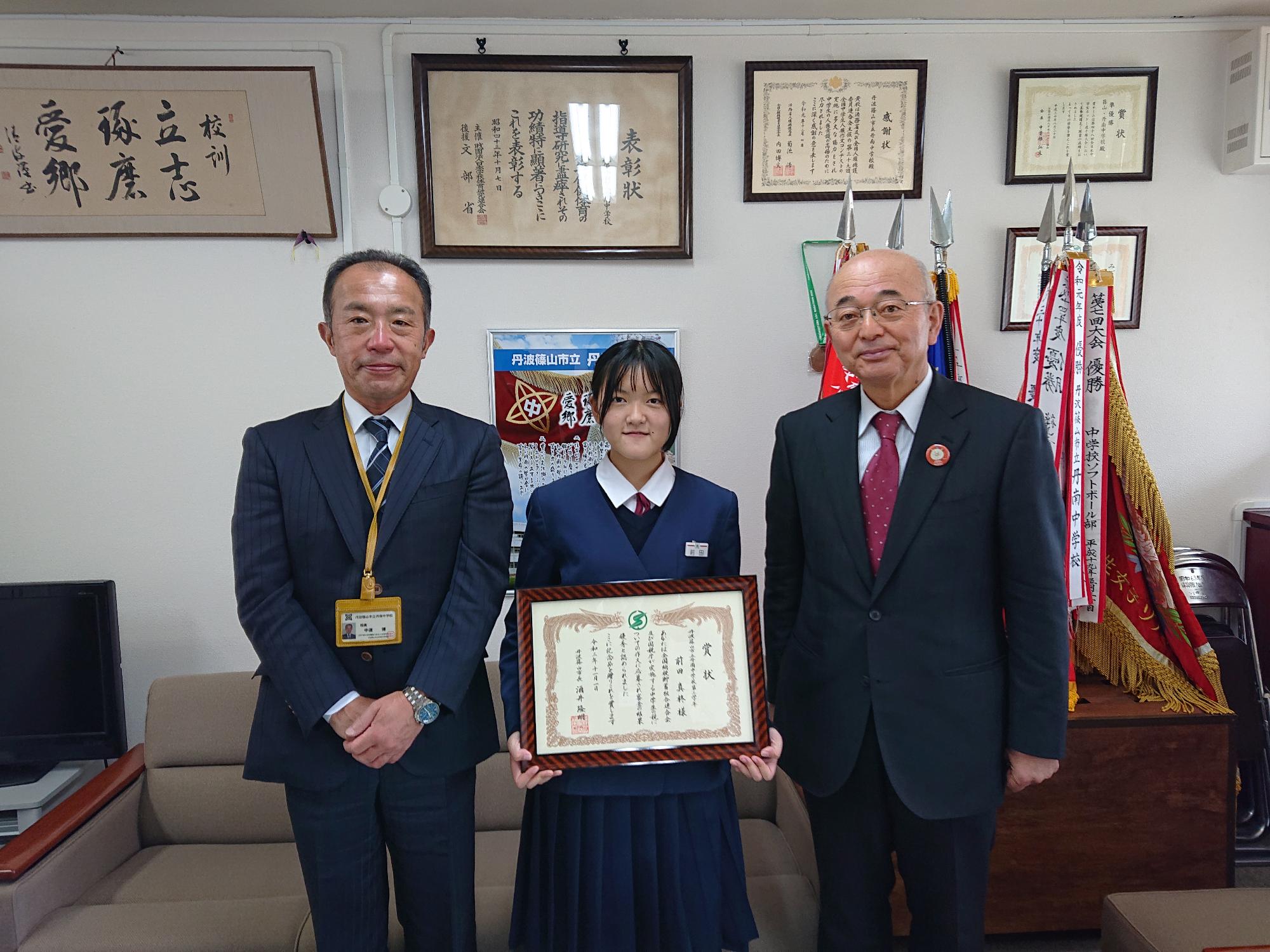 校長先生、市長、真ん中に額に入った表彰状を持った女の子が立っている