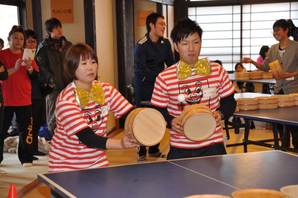 顔の書かれたTシャツに黄色の蝶ネクタイをつけた男女ペアの参加者が桶を持って卓球をしている写真